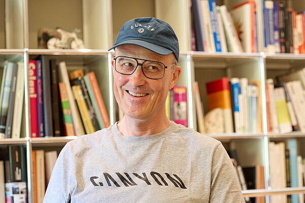 Ein Mann mit grauem T-Shirt mit der Aufschrift Canyon und blauem Baseball-Cap steht vor einem Bücherregal.