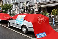 Ein Auto wird von einem roten Teppich bedeckt, der einen Radweg darstellt. Auf dem Auto steht ein Velo.