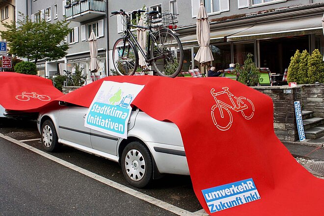 Mehr Bäume, mehr Platz für Velofahrerinnen und Fussgänger, das fordern verschiedene Organisationen. (Foto: ZVG) Ein Auto wird von einem roten Teppich bedeckt, der einen Radweg darstellt. Auf dem Auto steht ein Velo.