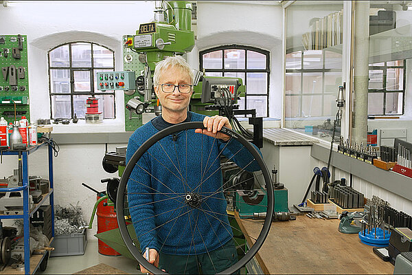 Ein Mann mit grauem Haar und blauem Pullover hält ein Fahrrad-Rad für Rennvelos in der Hand.