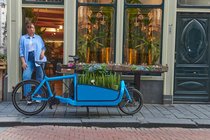 Steigende Kosten und Klimawandel motivieren zum E-Bike-Kauf. Eine Frau in Jeanshemd und Jeans steht hinter einem blauen Lastenrad. 