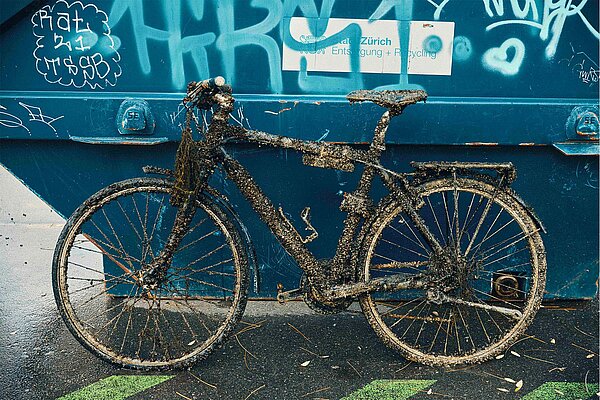 Aus der Limmat gefischtes von Muscheln bewachsenes Rostiges Velo steht vor blauem Züri Container