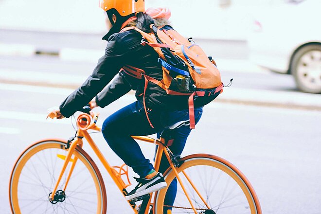 Eine Person fährt auf einem orangen Velo. Sie trägt einen orangen Rucksack und einen orangen Helm.