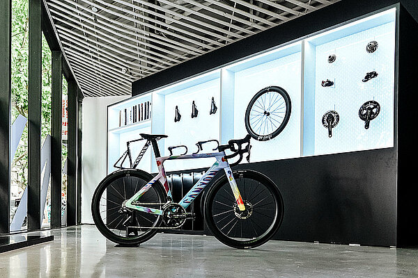 Ein Rennrad steht in einem Ausstellungsraum. Im Hintergrund hängen Helme, Laufräder und Fahrradzubehör.