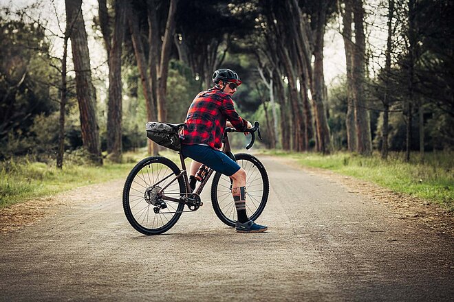 Ein Mann mit Carohemd sitzt auf dem Oberrohr eines Gravelbikes. 