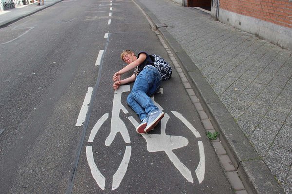 Ein junger Mann liegt am Boden auf einem Fahrradsignet, sodass es aussieht als würde er mit dem Velo fahren.