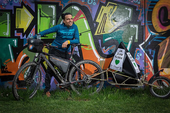 Michael Schoch alias Dr. Chopf mit Velo und Anhänger vor Graffitiwand.