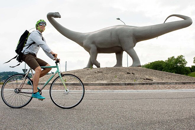 Ein Mann fährt auf einem Velo durch einen Kreisel, auf dem Kreisel steht ein Dinosaurier