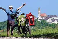 Ein Mann steht neben einem Tourenrad mit Packtaschen und hebt die Arme in die Luft.
