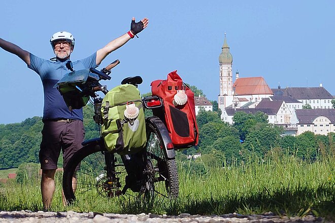 Ein Mann steht neben einem Tourenrad mit Packtaschen und hebt die Arme in die Luft.