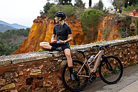 Moustache Dimanche Generation 3. Eine Rennradfahrerin sitzt auf einer Mauer neben ihrem Bike.