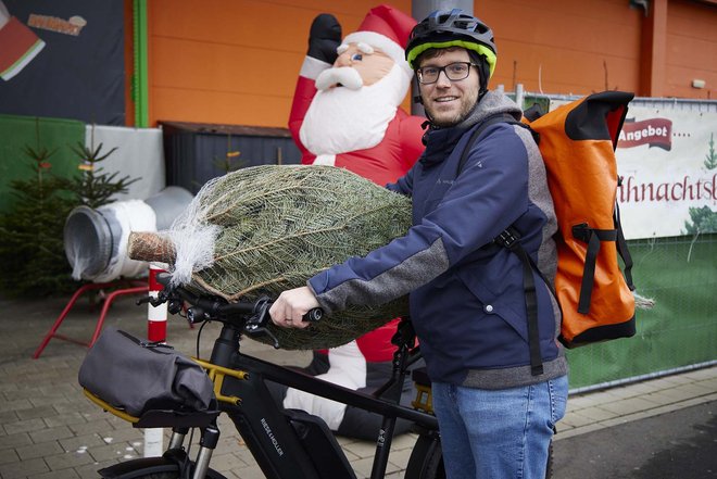 Wie man einen Weihnachtsbaum sicher mit dem Velo transportiert. Ein Mann schiebt ein Fahrrad, auf dem Lenker liegt ein Christbaum.