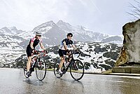 Eine Frau und ein Mann fahren mit Rennvelos auf einer Passstrasse. Im Hintergrund liegt Schnee.