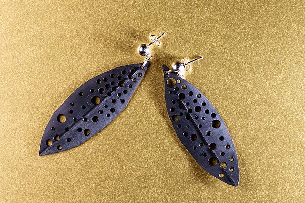 Blattförmige Ohrringe aus Veloschlauch mit Lochmuster auf goldenem Untergund.