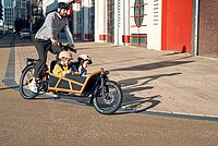 Riesemüller Cargobike fährt mit zwei Kindern vor roter Hauswand vorbei