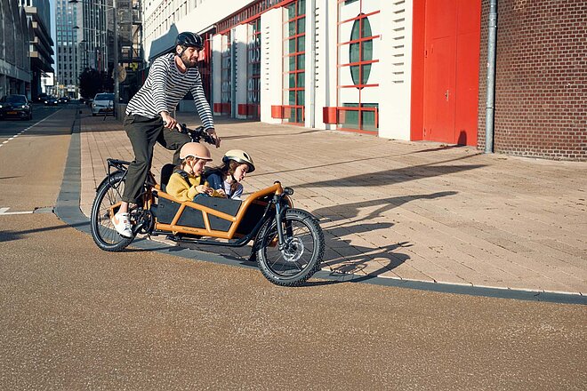 Riesemüller Cargobike fährt mit zwei Kindern vor roter Hauswand vorbei