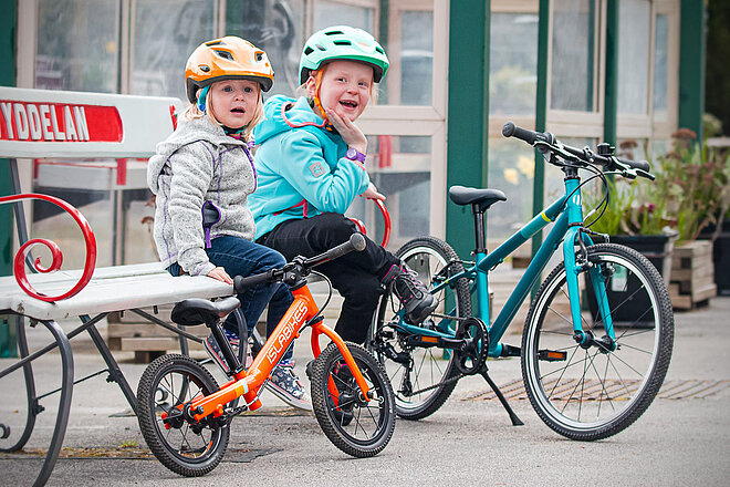 Kindervelo Marke Islabikes gibt auf. Zwei Kinder mit Fahrradhelmen sitzen auf einer Parkbank.