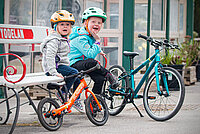 Kindervelo Marke Islabikes gibt auf. Zwei Kinder mit Fahrradhelmen sitzen auf einer Parkbank.