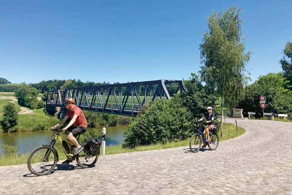 Locker durch die Schweiz radeln: Vier Routen zwischen Bodensee und Genfersee. Ein Mann und eine Frau auf E-Bikes, im Hintergrund ein Fluss.