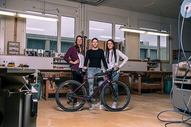 drei Frauen stehen hinter einem violetten Velo in einer Holzwerkstatt