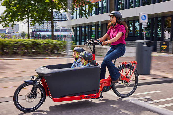 Trek Fetch Cargobikes. Eine Frau transportiert zwei Kinder in einem roten Lastenrad. 