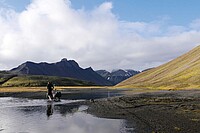 Ein Mann schiebt sein Fahrrad durch einen Fluss. Im Hintergrund hohe Berge