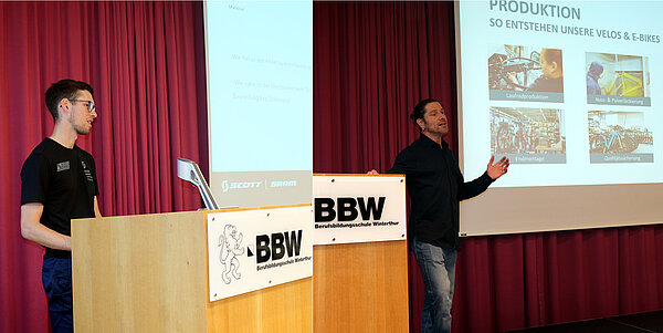 Zwei Männer sprechen auf einer Bühne zum Publikum.