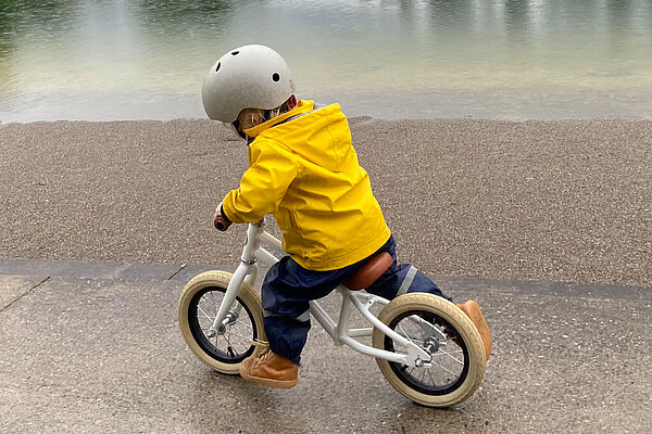 Kleinkind in gelber Regenjacke fährt auf dem Laufrad einem Seeufer entlang.