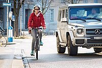 Eine Velofahrerin mit roter Jacke wird mit wenig Abstand von einem Auto überholt.