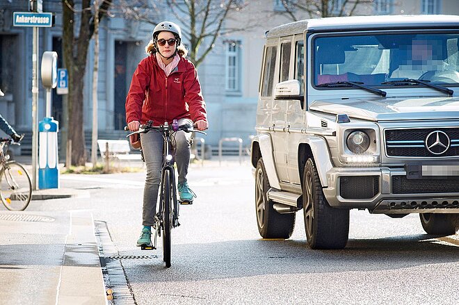 Eine Velofahrerin mit roter Jacke wird mit wenig Abstand von einem Auto überholt.