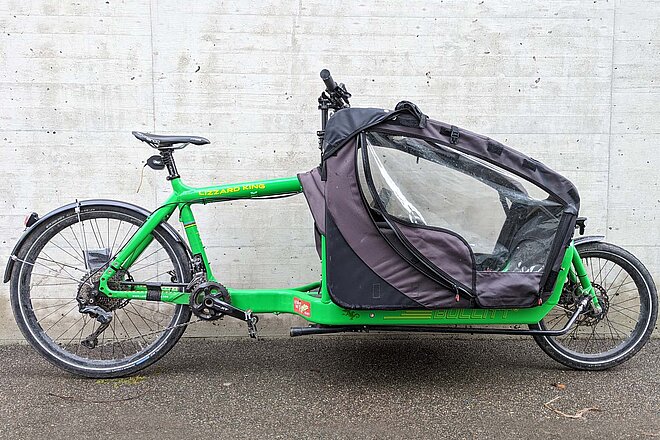 Bullitt Cargobike Test. Ein grünes Lastenrad steht vor einer Betonwand.