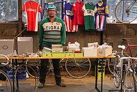 Ein Mann mit Fahrracap und Vintage-Shirt steht hinter einem Verkaufstisch mit Fahrradteilen.