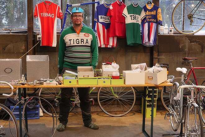 Ein Mann mit Fahrracap und Vintage-Shirt steht hinter einem Verkaufstisch mit Fahrradteilen.