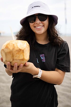 Samantha Soriano. Eine junge Frau mit weissem Hut hält eine Kokosnuss in die Kamera