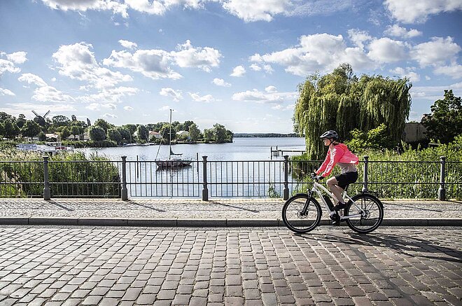Eine junge Frau fährt auf einem Pedelec über eine Brücke. in Hintergrund sind ein See und blauer Himmel mit Wolken zu sehen.