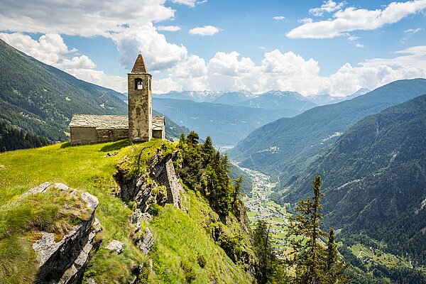 Alpine Landschaft mit Steinkirche auf einem Hügel. Im Hintergrund Berge.
