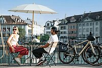 Eine Frau und ein Mann sitzen an einem Bistrotisch am Ufer eines Flusses.