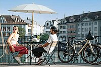Eine Frau und ein Mann sitzen an einem Bistrotisch am Ufer eines Flusses.