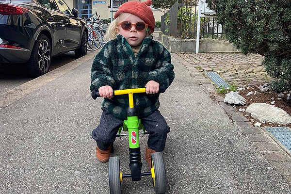 Kleinkind auf vierräderigem grünen Velo mit Sonnbrille und Karofleece