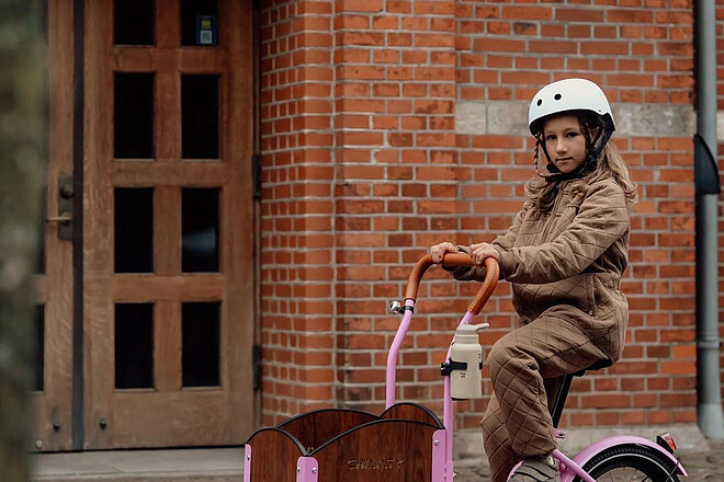 Mädchen in beigem Overall auf pinkem Kinder Lastenrad vor Backsteinmauer