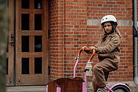 Mädchen in beigem Overall auf pinkem Kinder Lastenrad vor Backsteinmauer