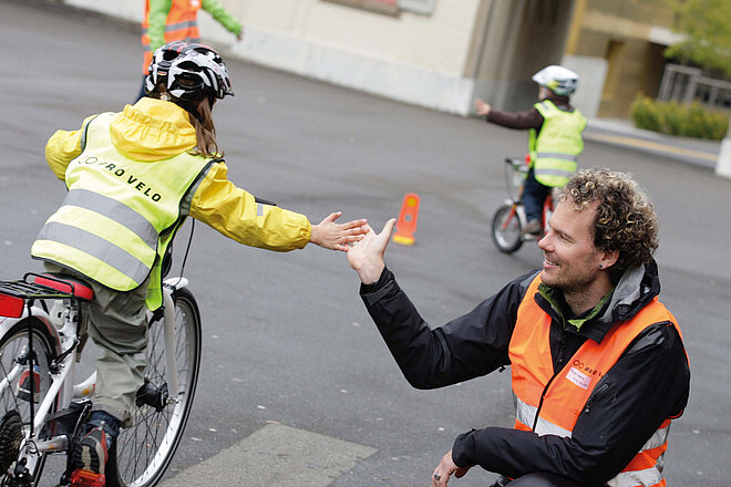 Ein Kind in gelber Jacke und Leuchtweste fährt mit dem Velo auf einem Pausenplatz