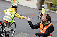 Ein Kind in gelber Jacke und Leuchtweste fährt mit dem Velo auf einem Pausenplatz