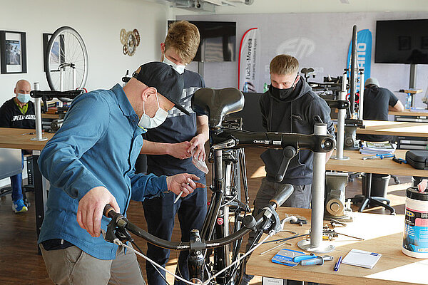 Kurs als Velomechaniker Quereinsteiger. Drei Männer mit Nasen-Mund-Schutz arbeiten an einem Fahrrad. 
