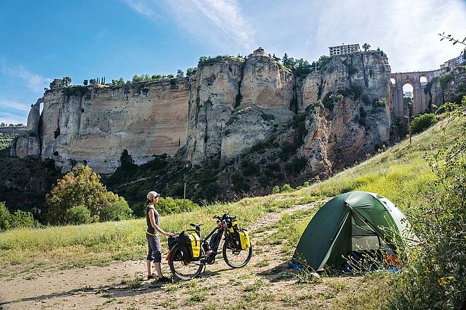 Die erste «wilde» Nacht vor den Toren der historischen Stadt Ronda. Im Mai 2021 schwingt sich die Journalistin Susanne Brüsch in Andalusien auf den Sattel ihres E-Bikes und «surft» auf der Öffnungswelle nach dem Corona-Lockdown vom Atlantik bis an den Rhein. Eine Frau steht auf einer Wiese vor ihrem Zelt.