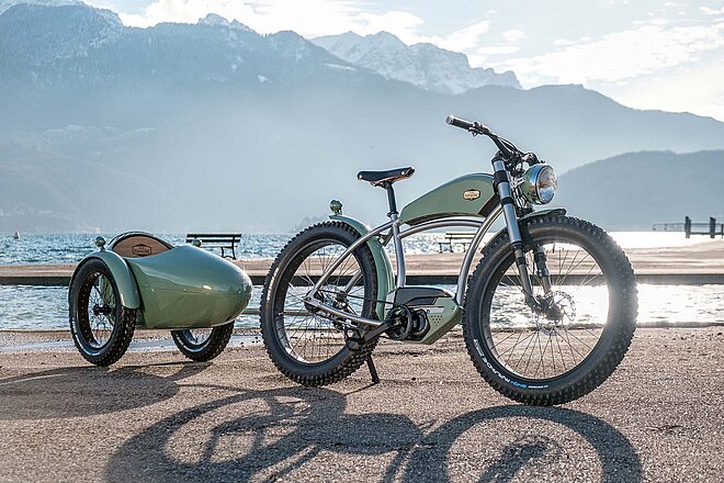 Ein Gespann, wie es in der Velowelt derzeit seinesgleichen sucht: «Origine» mit Sidecar. Ateliers Heritage Bike. Vintage E-Bike mit Anhänger steht vor einem See.