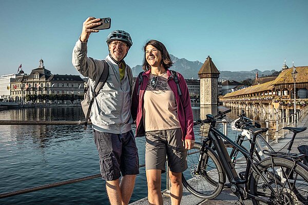 Route 1291. Die beste E-Bike Tour der Schweiz. Ein Mann und eine Frau stehen vor der Kappelbrücke in Luzern und machen ein Selfie.