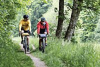 Eine Frau und ein Mann fahren mit Gravelbikes auf einem Feldweg durch eine gürne Landschaft.