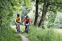 Eine Frau und ein Mann fahren mit Gravelbikes auf einem Feldweg durch eine gürne Landschaft.
