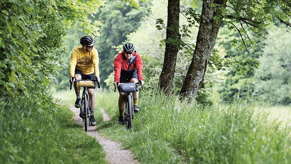 Eine Frau und ein Mann fahren mit Gravelbikes auf einem Feldweg durch eine gürne Landschaft.